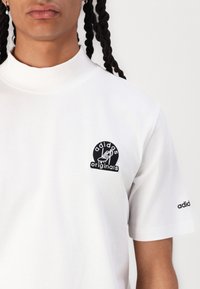 Tricou alb din bumbac cu guler înalt, având un logo brodat Adidas Originals de culoare neagră pe pieptul din stânga și un logo pe mânecă.