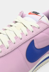 Κοντινό πλάνο ροζ αθλητικού παπουτσιού Nike με άσπρα κορδόνια, μπλε λογότυπο swoosh και ορατή ετικέτα γλώσσας που διαθέτει κόκκινη και μαύρη σήμανση Nike.