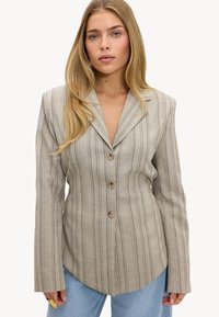 Femme aux longs cheveux blonds porte un blazer beige à fines rayures avec trois boutons et un jean bleu clair sur un fond blanc uni.
