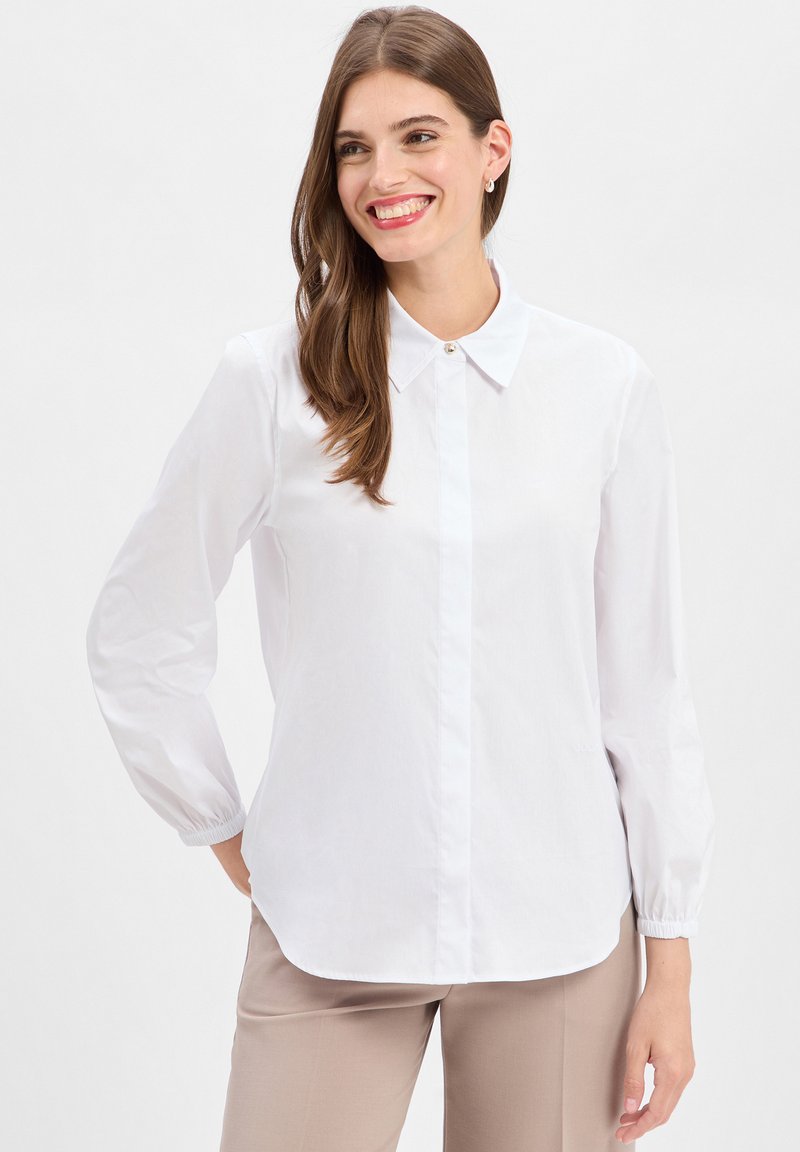 Vrouw die glimlacht, gekleed in een frisse witte blouse met lange mouwen en beige broek tegen een effen achtergrond.