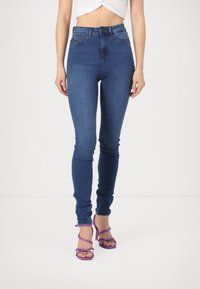 Jean bleu taille haute, coupe ajustée, porté avec un top blanc court et des sandales à talons avec lanières violettes sur fond blanc.
