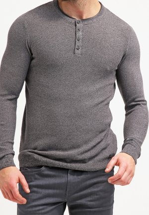 Hombre con suéter henley gris ajustado de manga larga con tres botones, combinado con pantalones gris oscuro, de pie frente a un fondo liso.