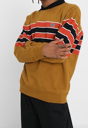 Osoba ubrana w musztardową bluzę z czarnymi, czerwonymi i białymi poziomymi paskami oraz logo "obey" na piersi, z rękami skrzyżowanymi na klatce piersiowej.