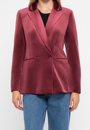 Kobieta ubrana w dopasowaną, dwurzędową bordową welurową marynarkę, czarny top i niebieskie dżinsy, stojąca na jednolitym tle.