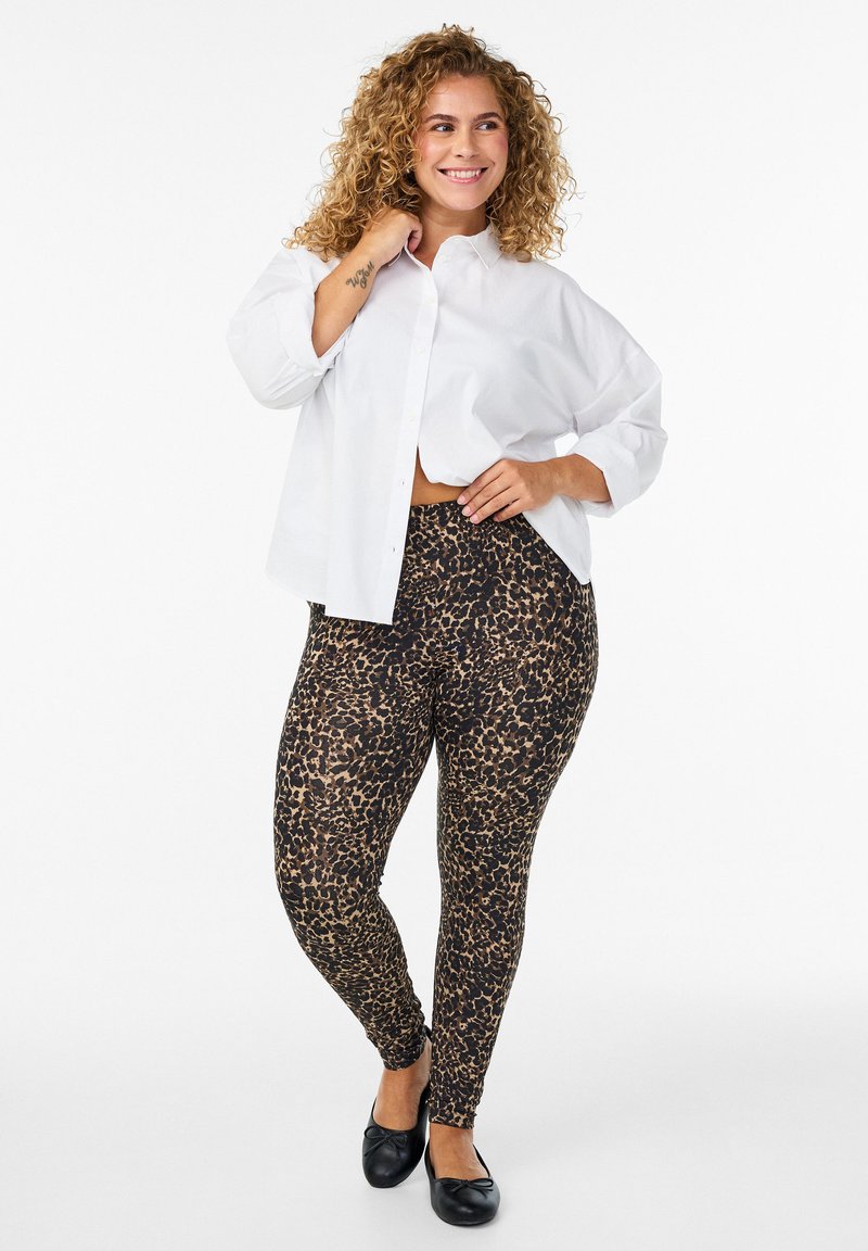 Hvid skjorte med knapper, leopardmønstrede leggings i brune og sorte farver, tekstureret stof og sorte flade sko. Modellen poserer med den ene hånd på hoften.
