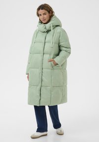 Kobieta w długim, jasnoniebieskim płaszczu puchowym z dużym kapturem, ciemnoniebieskich dżinsach i białych trampkach, stojąca z jedną ręką w kieszeni.