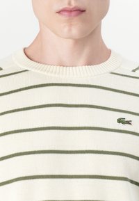 Pulover crem cu dungi orizontale verzi, guler ribbit și logo-ul brodat cu crocodil pe partea stângă a pieptului. Material texturat din tricot.