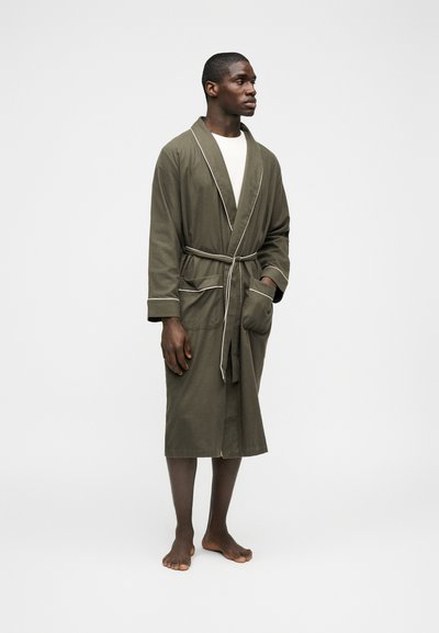 Άνδρας που στέκεται ξυπόλητος, φοράει ρόμπα ελιάς με λευκή επένδυση, δεμένη στη μέση πάνω από λευκό πουκάμισο, με φόντο ένα απλό λευκό φόντο.