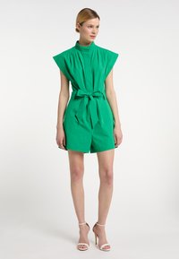 Salopetă verde din bumbac, fără mâneci, cu guler înalt, umeri largi și talie legată. Prezintă pantaloni scurți lejeri și o textură netedă.