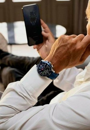 Montre connectée avec un bracelet en silicone bleu, un cadran noir rond, un écran numérique et des fonctionnalités multi-usages, tenue près d'un smartphone.