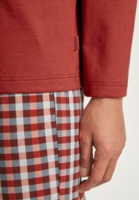 Chemise rouge à manches longues en matériau texturé, associée à un pantalon à carreaux rouge, blanc et bleu avec un motif en chevron. Étiquette visible sur la chemise.