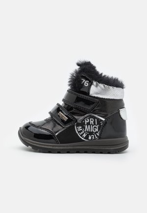 Bota de invierno para niños en negro y plata, con borde de piel sintética, dos correas de Velcro y un parche circular con texto blanco en el lateral.