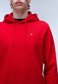 Sudadera roja con cordón, que presenta un pequeño logo texturizado en el pecho. Tejido suave, corte relajado y mangas largas.