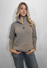 Pullover lavorato a maglia color beige chiaro con colletto alto, dettagli in bottoni e polsini a coste. Indossato con jeans in denim blu.