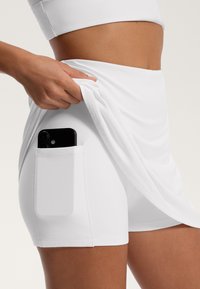 Beli športni šort, ki ima stransko žep, v katerem je pametni telefon, izdelan iz mehkega, raztegljivega materiala, z oblikovanim dizajnom in gladko teksturo.
