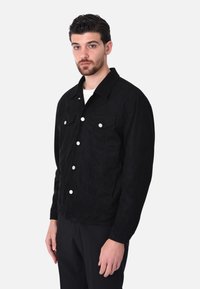 Giacca in suede nera con bottoni argentati, due tasche sul petto e colletto. Presenta un design classico con cuciture visibili.