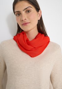 Femme aux longs cheveux bruns et taches de rousseur porte un pull beige et une écharpe rouge vif enroulée lâchement autour du cou.