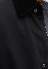 Nahaufnahme einer schwarzen Jacke mit einem Cordkragen, Metall-Druckknöpfen und einem teilweise sichtbaren Reißverschluss auf der rechten Seite.