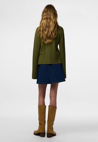 Olivno zelena pletena majica, modra denim krilo v črki A in rumeni škornji do kolen z teksturirano površino in lesenimi petami, prikazano od zadaj.