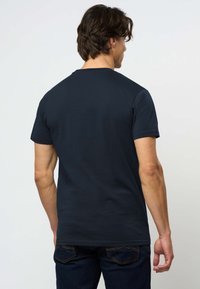 Muškarac s tamnom kosom, obučen u običnu crnu majicu kratkih rukava i tamne traperice, okrenut leđima prema jednostavnoj svijetloj pozadini.