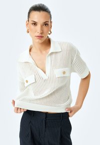 Polo shirt bianco a maglia con maniche corte, caratterizzato da una trama a trama aperta, due tasche sul petto e dettagli con bottoni in legno.