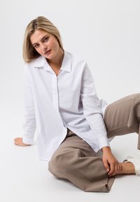 White button-up shirt with long sleeves, relaxed fit, and a classic collar, paired with beige trousers, on a light background.