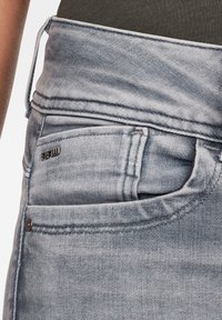 Jeans de mezclilla azul claro deslavados con cintura alta, lazo de cinturón visible, bolsillo frontal y etiqueta de metal con el logo "RAW" en el borde del bolsillo.