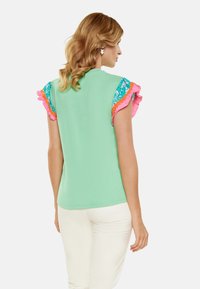 Top verde mentă cu mâneci din volane cu model roz și albastru și o textură netedă. Stilizat cu pantaloni albi, văzut din spate.