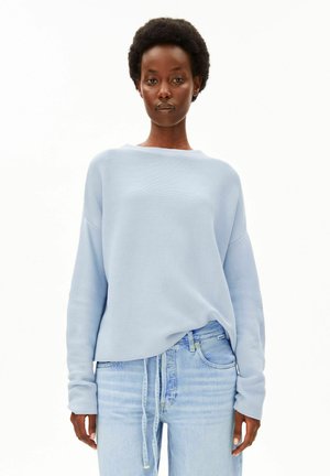 Femme aux cheveux afro courts portant un pull bleu clair à manches longues et un jean bleu clair sur un fond blanc uni.