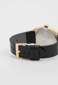 Black leather watch strap with a gold metal buckle and hexagonal gold case, featuring a clear glass back and embossed branding.