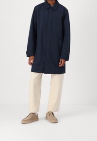 Persoon in een marineblauwe middellange jas, beige broek, witte sokken en lichtbruine casual schoenen staat tegen een effen achtergrond.