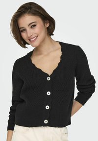 Black knitted cardigan with a scalloped V-neckline, featuring five large white buttons and ribbed cuffs. Textured pattern throughout.