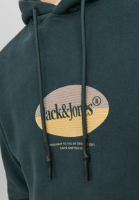 Sudadera verde oscura con un gran gráfico circular que presenta "Jack & Jones" en texto negro y un patrón de madera. Cuerdas con puntas negras.