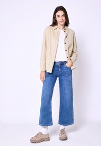 Beige corduroy button-up shirt over a white T-shirt, paired with wide-leg blue jeans and beige shoes, standing against a white background.