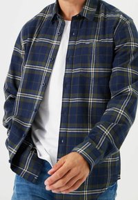 Mörkblå och grön rutigt flanellskjorta med vita detaljer, knäppning fram, krage och en enkel bröstficka. Buren över en vit t-shirt.