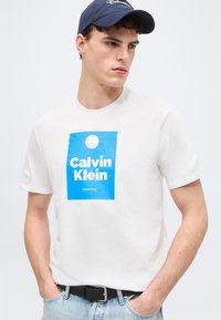 Νεαρός άνδρας που φοράει ένα λευκό μπλουζάκι Calvin Klein με μπλε λογότυπο και μια ναυτική καπέλο, στέκεται με τα χέρια στις τσέπες κοιτώντας προς τα αριστερά.