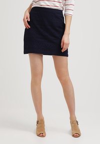 Mini-jupe bleu marine en tissu texturé avec des découpes horizontales et une taille ajustée, associée à des bottines ouvertes à bout découvert couleur beige clair.