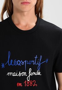 Svart bomullst-shirt med flerfärgad text: blå "le coq sportif," vit "maison fondée," och röd "en 1882." Klassisk rund hals.