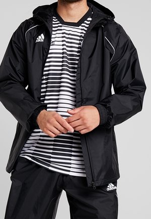 Mann trägt schwarze Adidas-Jacke und Hose mit schwarz-weiß gestreiftem Hemd vor einfarbigem Hintergrund.
