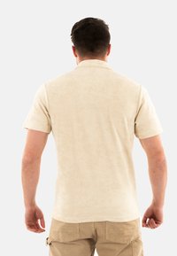 Polo shirt beige a maniche corte con una texture morbida, caratterizzato da un colletto standard e una vestibilità rilassata. La parte posteriore è liscia e senza decorazioni visibili.