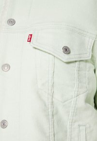 Giacca in velluto a coste verde chiaro con bottoni a pressione argentati, grandi tasche frontali e un'etichetta rossa Levi's sulla tasca del petto. Dettaglio del tessuto testurizzato visibile.