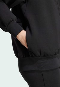 Black zip-up jacket with puffed sleeves and ribbed cuffs. Features a side pocket and elastic hem. Smooth fabric texture.