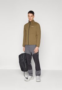 Olive green zip-up jacket with a black zipper and logo, paired with grey and black tapered pants. The model holds a black backpack.