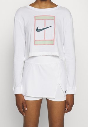 Persoon draagt een wit, crop top met lange mouwen en een Nike-logo en een tennisbaanafbeelding, gecombineerd met witte korte rok met Nike-logo.