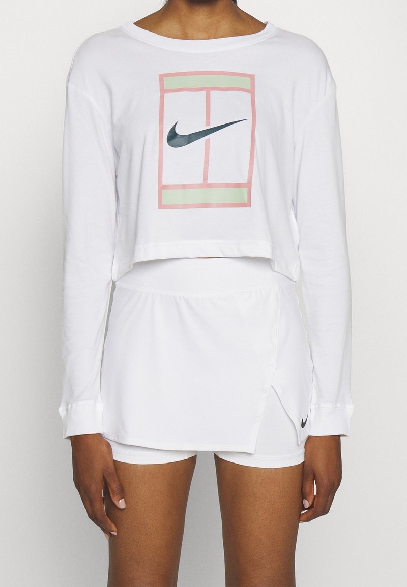Personne portant un crop top à manches longues blanc avec le logo Nike et un motif de court de tennis, assorti à un short-jupe blanc avec le logo Nike.