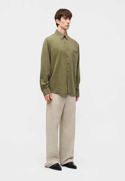 Νεαρός άνδρας στέκεται φορώντας ένα φαρδύ πουκάμισο υπερμεγέθους σε χακί απόχρωση με κουμπιά, μπεζ παντελόνι φαρδιάς γραμμής και μαύρα μυτερά παπούτσια, σε λευκό φόντο.