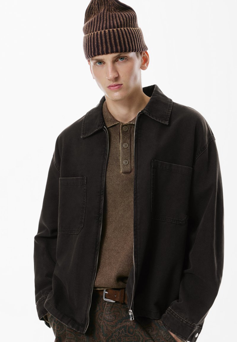 Giacca di jeans nera con frontale bottonato, due tasche sul petto, indossata sopra una maglietta polo marrone a trama. Berretto a coste marrone sulla testa.
