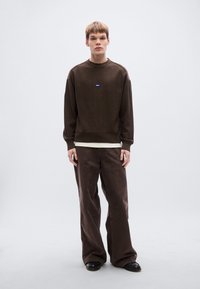 Pull en maille marron avec ourlet et poignets côtelés, arborant un petit logo bleu. Associé à un pantalon marron à jambes larges et des chaussures noires.