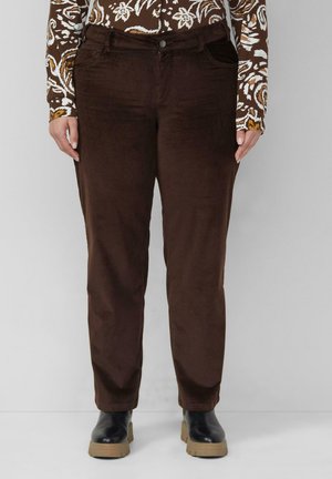 Personne portant un pantalon en velours côtelé marron foncé, une chemise à manches longues marron et blanche à motifs, et des bottes noires avec des semelles couleur beige, debout devant un fond gris.