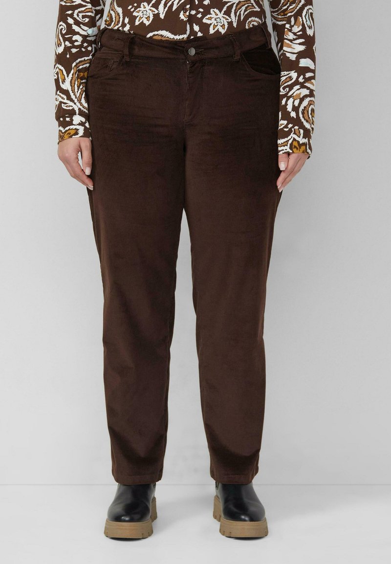 Personne portant un pantalon en velours côtelé marron foncé, une chemise à manches longues marron et blanche à motifs, et des bottes noires avec des semelles couleur beige, debout devant un fond gris.